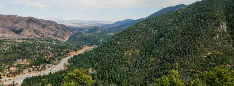 Mountain valley covered in dense evergreen forest with a highway winding through the landscape and distant plains visible on the horizon.